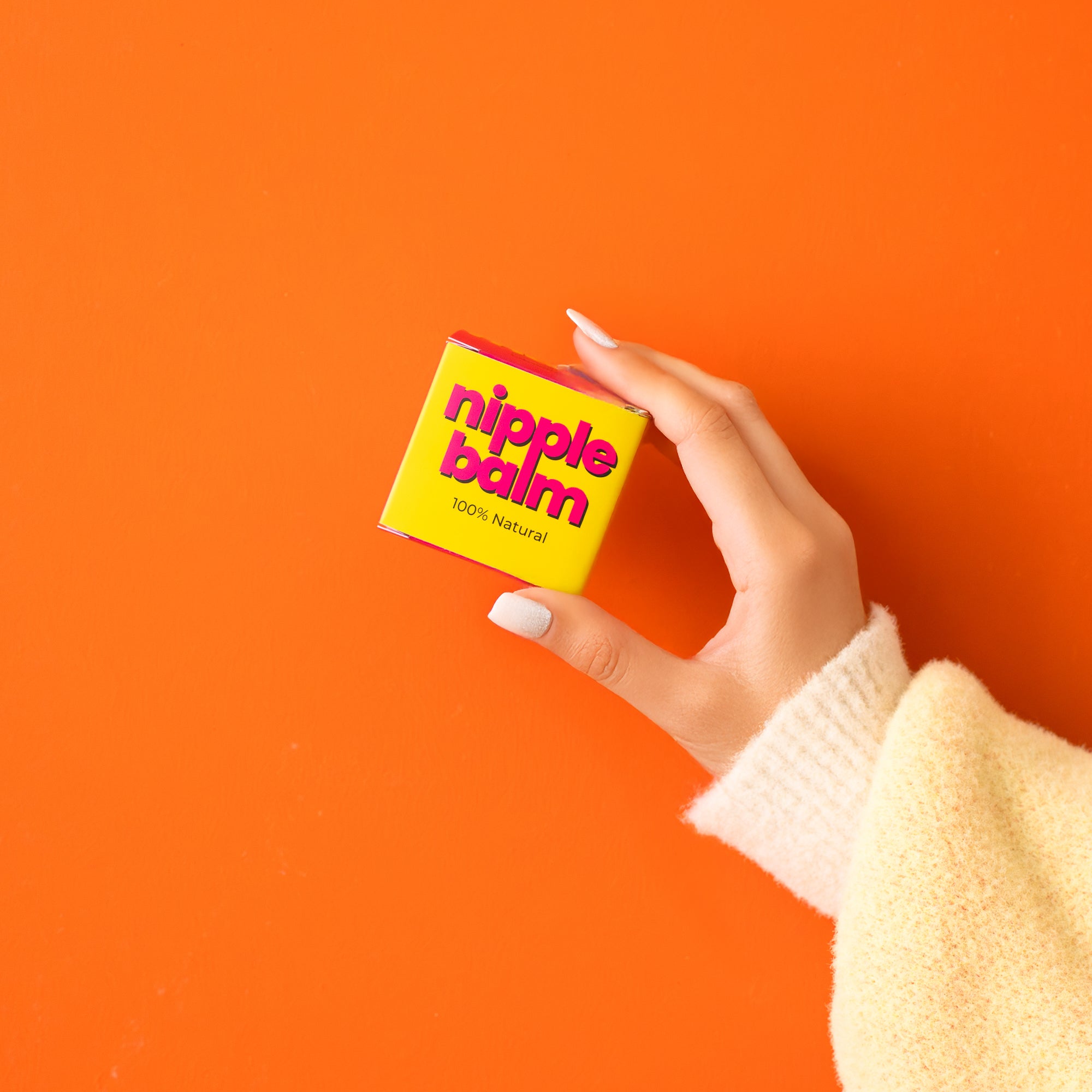 Hand holding a small box labeled 'nipple balm' against an orange background
